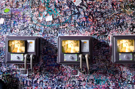 A highly piexlated image of dense and hard-to-read graffiti on a wall, with three very old looking televisions mounted on the same wall 
