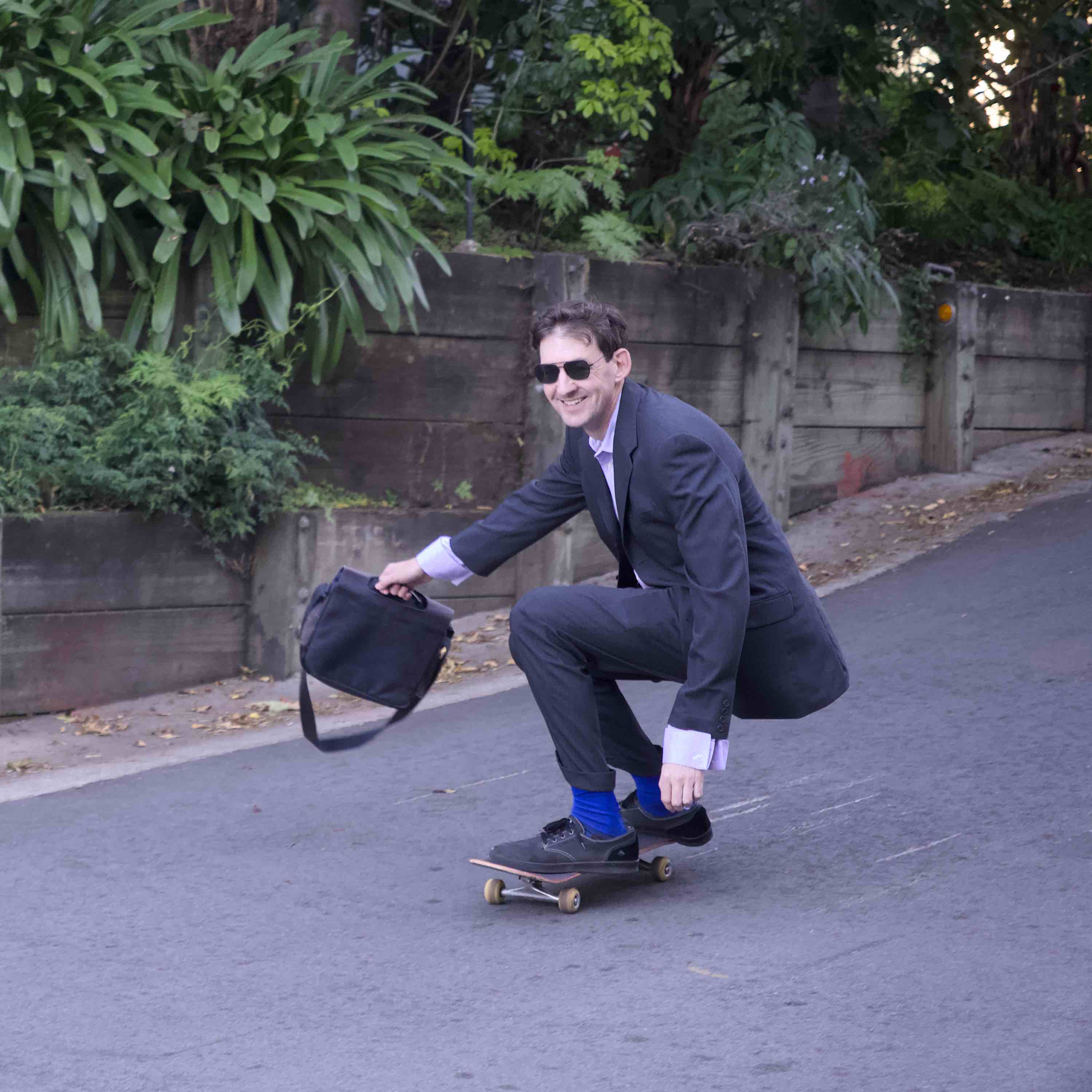 A picture of Chris Potts skateboarding down a hill in a business suit holding a messenger bag in his right hand, looking very cool and smiling